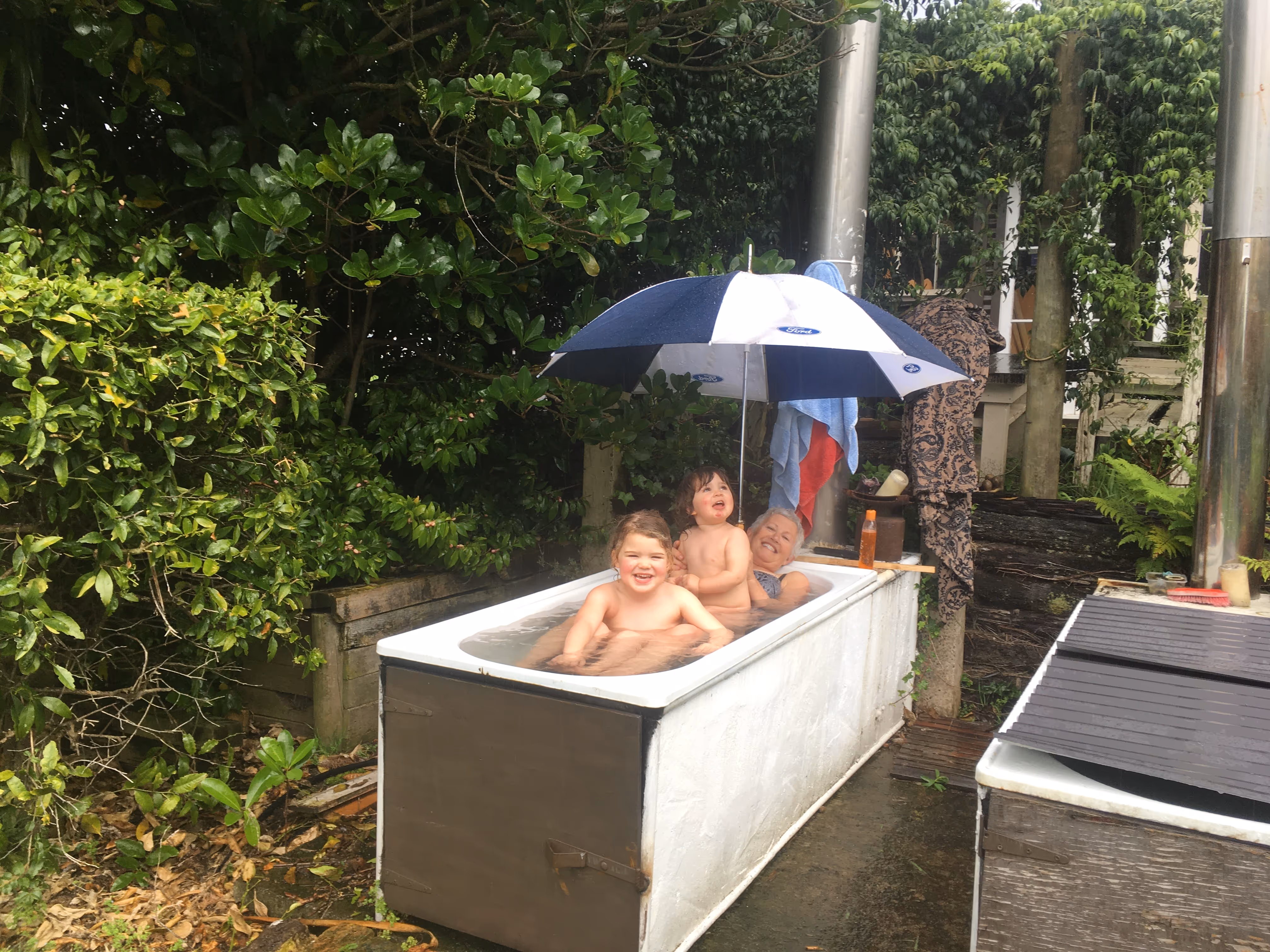 Two children and an older woman smiling while sitting in an outdoor bathtub with a Ford-branded umbrella overhead, surrounded by greenery, the inspiration behind Star Baths.