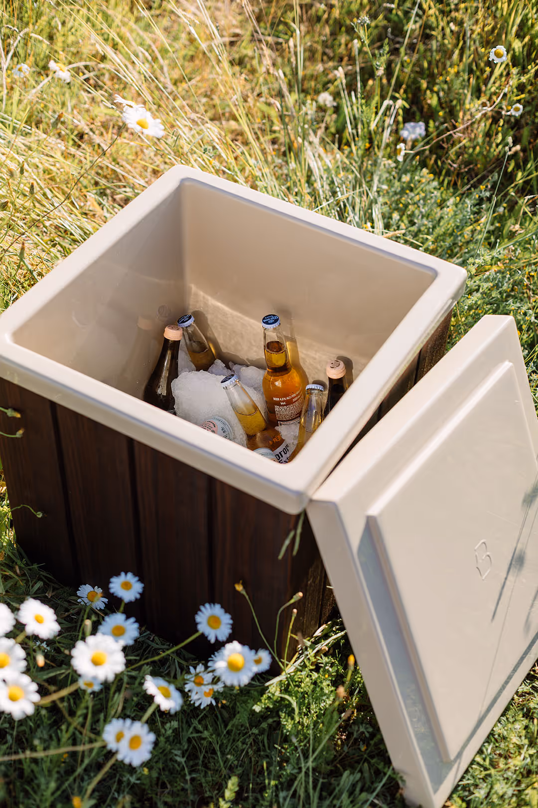 Star Baths insulated bathside storage box open to reveal chilled drinks on ice, set outdoors among daisies and summer grass.