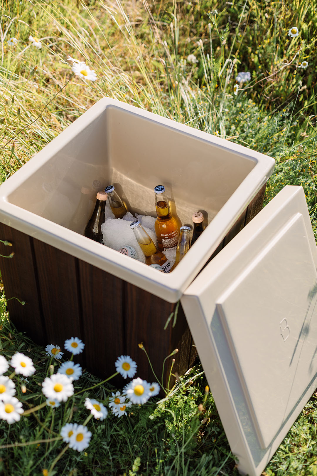 Star Baths insulated bathside storage box open to reveal chilled drinks on ice, set outdoors among daisies and summer grass.
