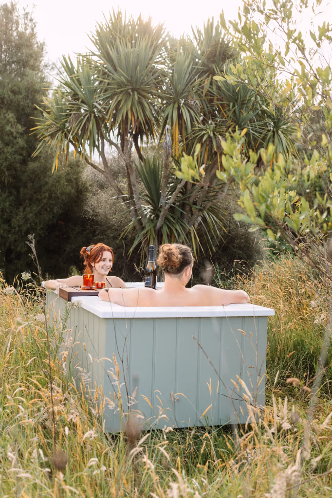 Couple relaxing together in a sage green Star Baths outdoor bath surrounded by native NZ bush, with wine and Retreat Tray
