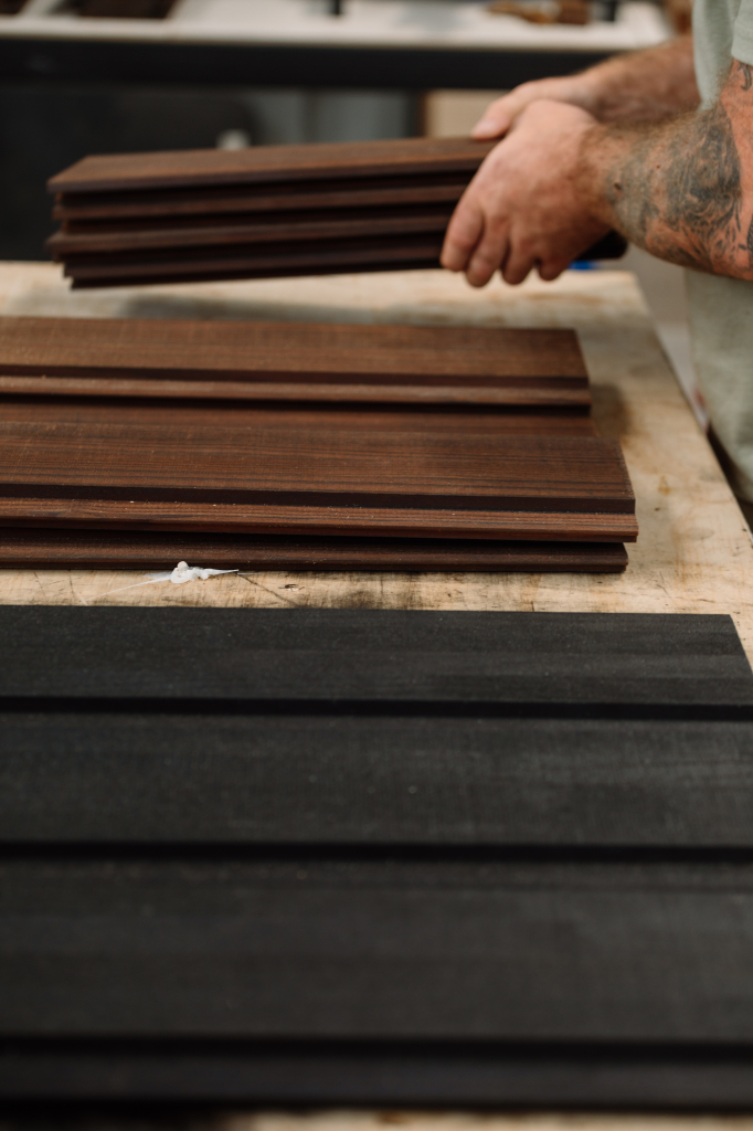 Close-up of a person holding stacked dark wooden planks with other brown and black planks on a workbench being prepared for a Star Bath.