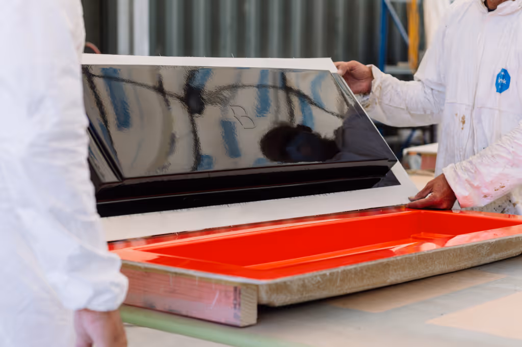 Two Star Baths team, in white protective suits,  lifting a freshly moulded fibreglass panel from a bright red casting tray in the workshop.
