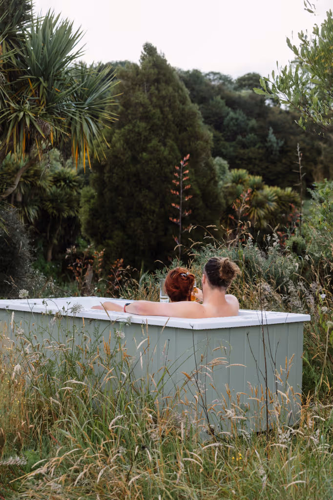 Couple relaxing together in a sage green Star Baths outdoor bath surrounded by native New Zealand bush, cabbage trees and wild grasses