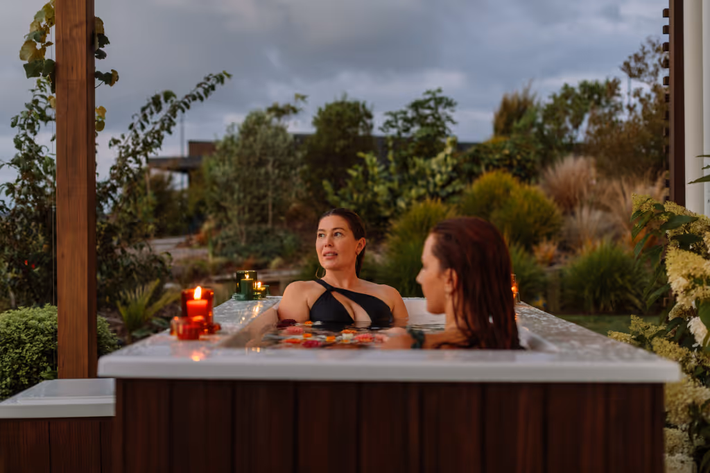 Two women relaxing together in a Star Baths outdoor bath at dusk, with floating flowers and candles, set within a landscaped NZ garden with timber pergola