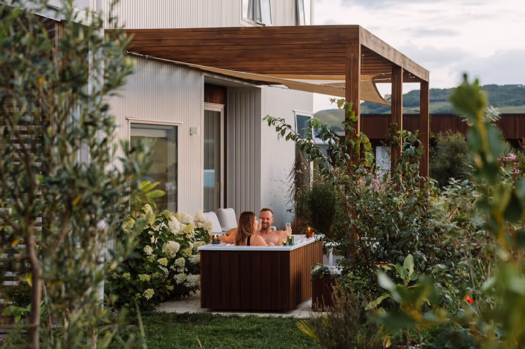 A couple relaxing in a timber-clad outdoor bath surrounded by greenery next to a modern house with a wooden pergola.