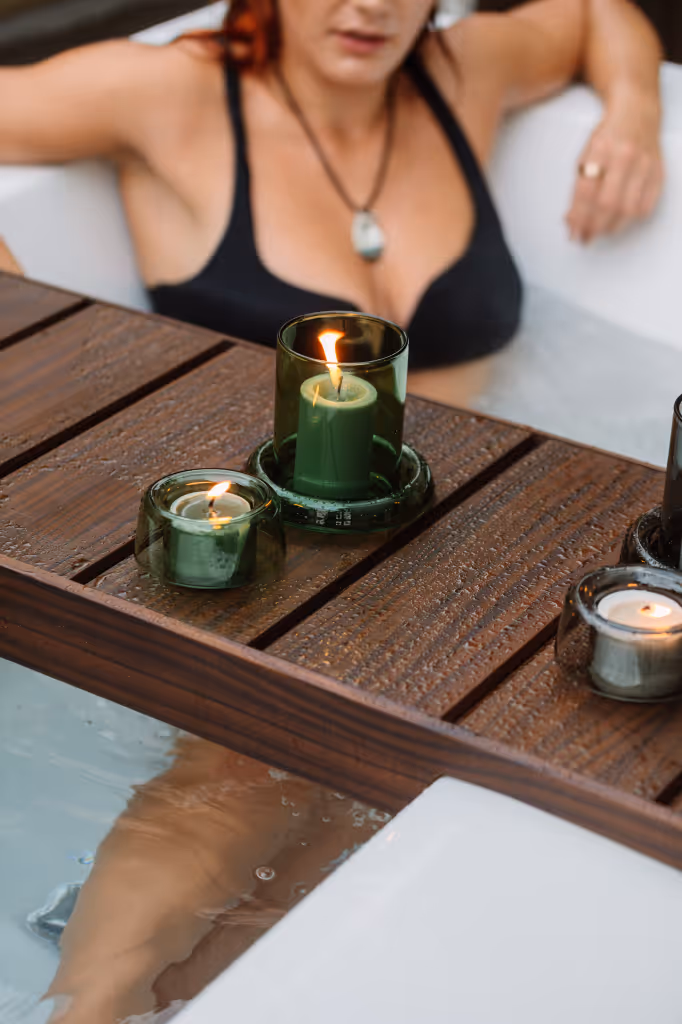 Woman in black bikini relaxing in an outdoor bath with a Star Baths timber Retreat Tray resting across the bath edge holding lit candles, creating an ambient evening bathing ritual