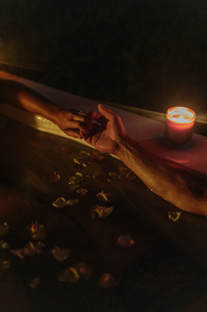 Two people holding hands in a candlelit bathtub filled with flower petals.