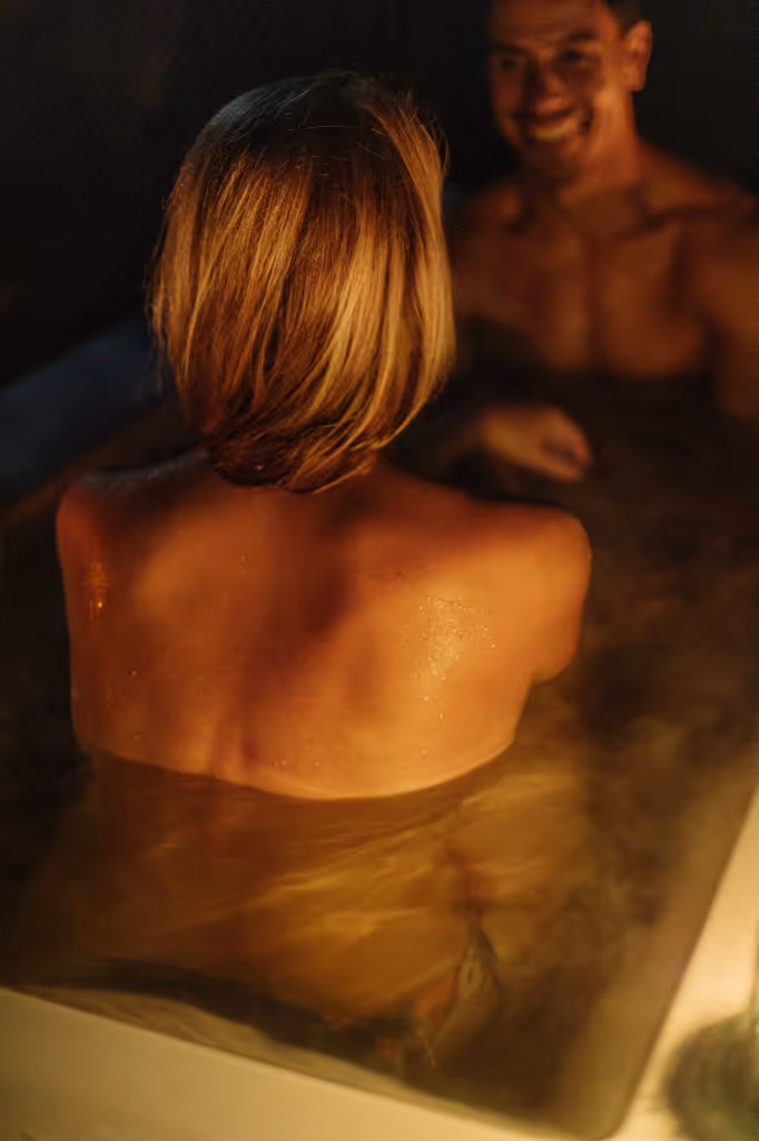 Man and woman relaxing in a steamy outdoor bath at night, the woman seen from behind with wet skin and blonde hair.