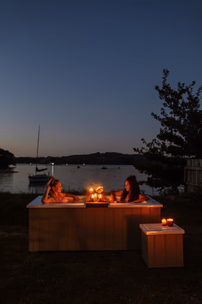 Two women relaxing in a candlelit Star Bath outdoors at dusk, overlooking Raglan Harbour with sailboats in the background and Bathside Storage beside.