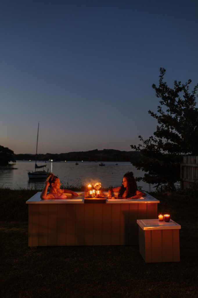 Two women relaxing in a candlelit Star Bath outdoors at dusk, overlooking Raglan Harbour with sailboats in the background and Bathside Storage beside.