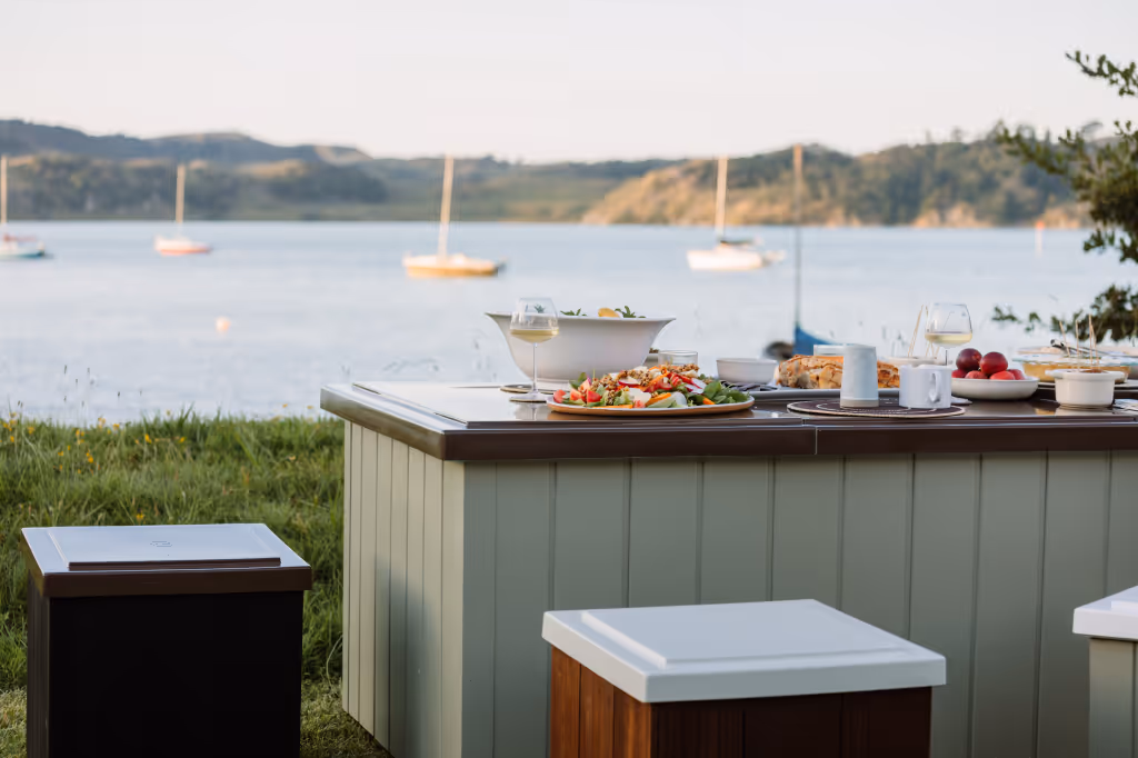 StarBath and lid as an outdoor dining table with various dishes and glasses of white wine by a harbour with sailboats and hills in the background.