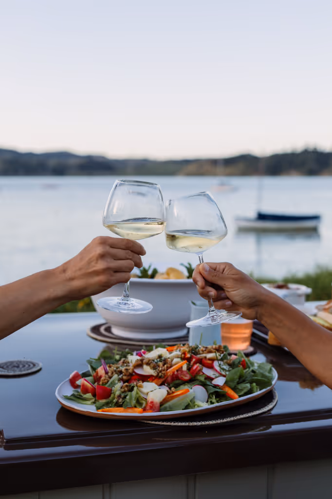 Two people toasting white wine over a Star Baths Eclipse Lid used as an outdoor dining table, Raglan harbour and sailboats in background