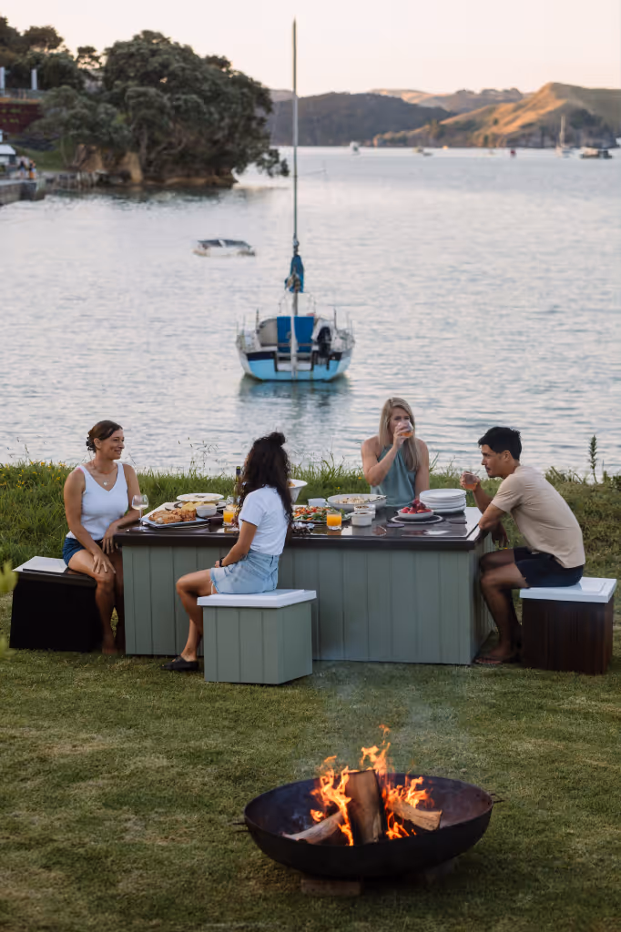 Four friends sitting outdoors around a Star Bath Eclipse fitted lid as a table with food and drinks with bathside storage as seating, overlooking a New Zealand harbour at sunset, with a fire pit in the foreground.