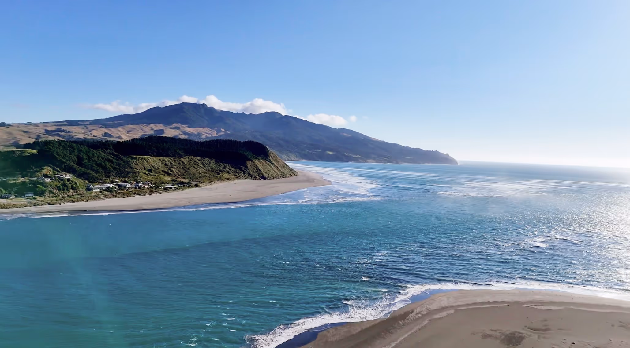 Aerial drone view of a Raglan, New Zealand harbour entrance, coastline with sandy beaches, blue ocean waves, and green hills under a clear blue sky.