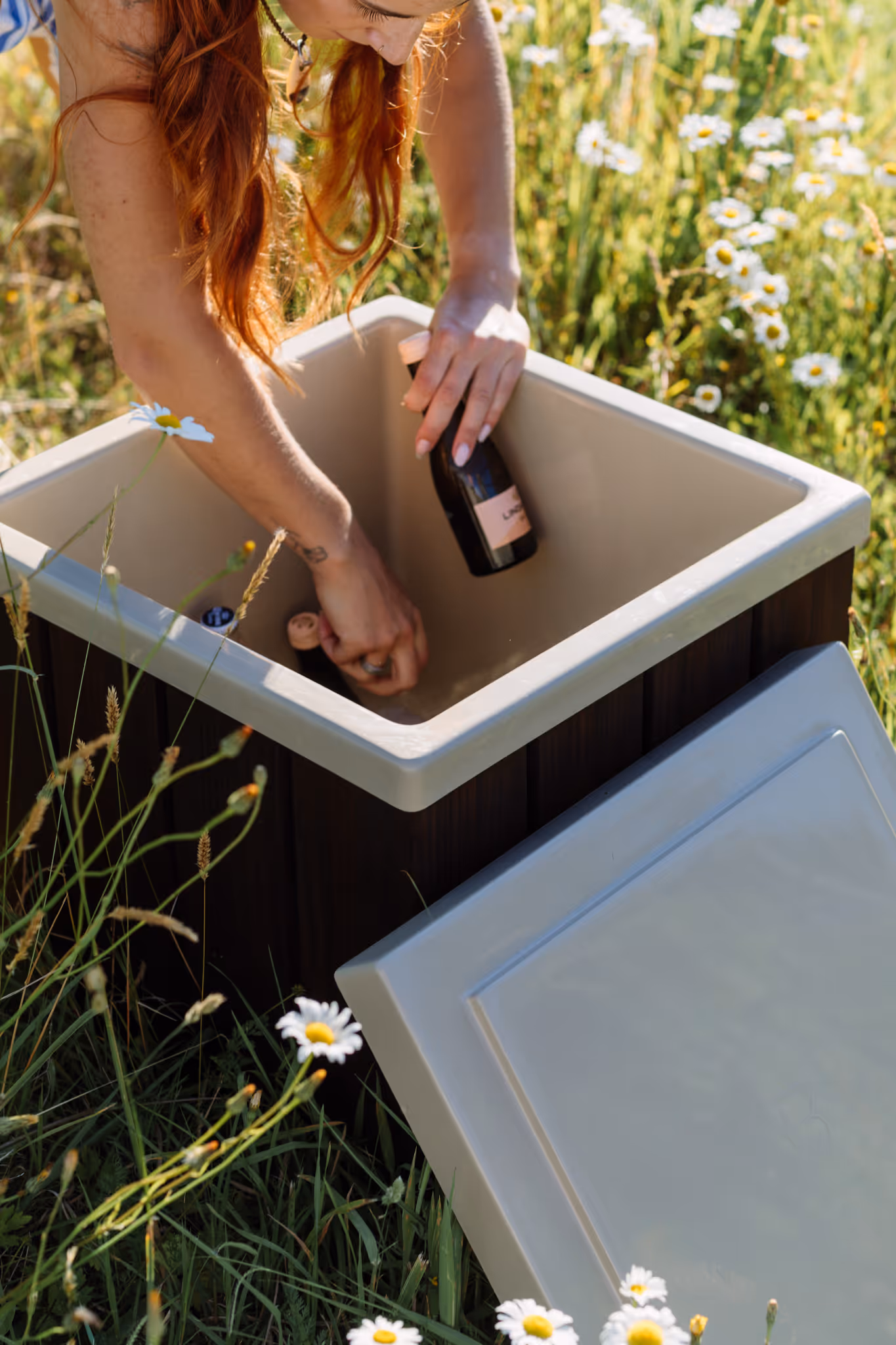 Woman reaching into a Star Baths insulated storage box to retrieve a wine bottle, surrounded by wildflowers — perfect outdoor entertaining companion