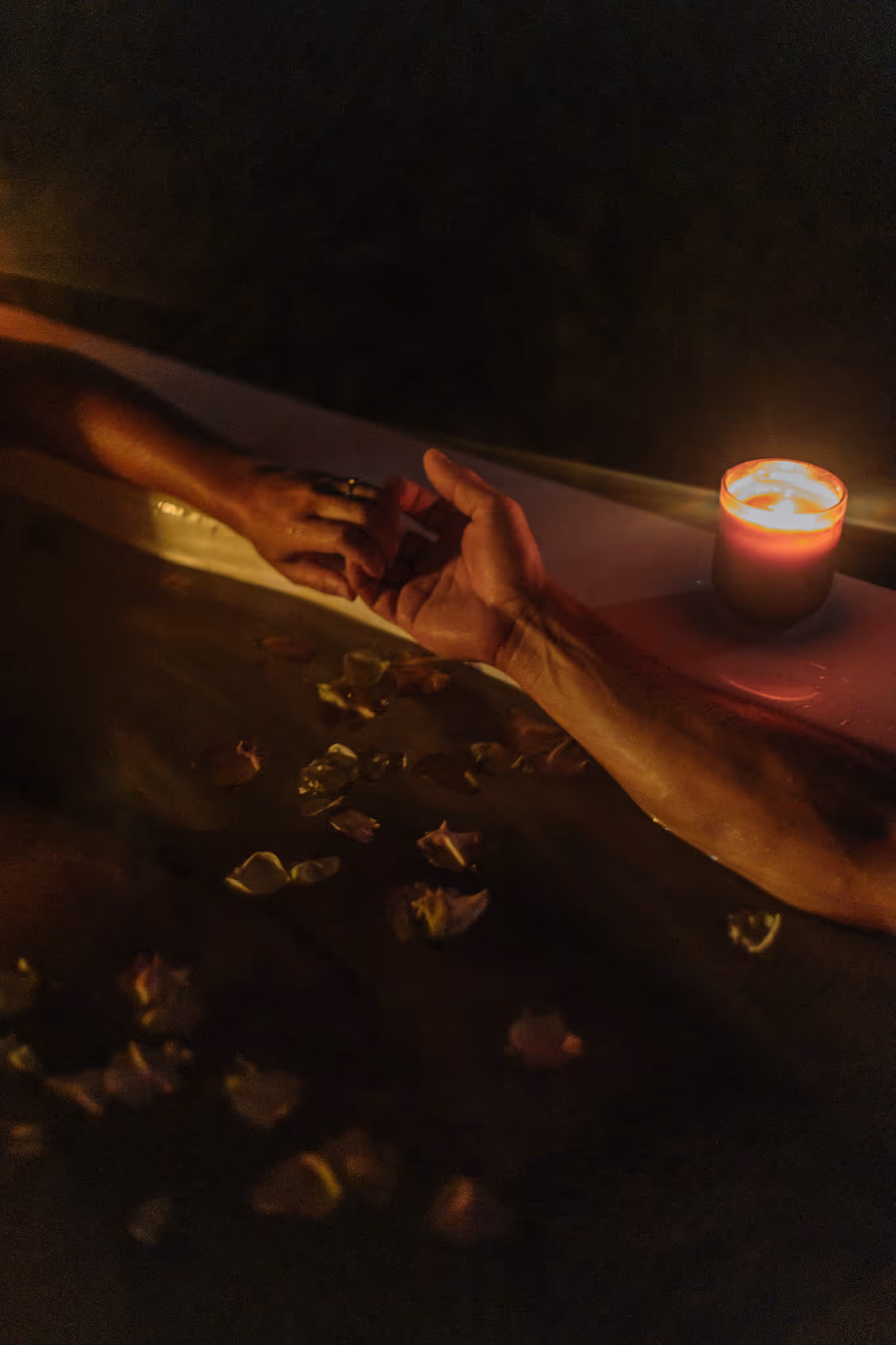 Two hands reaching toward each other across a Star Baths outdoor bath at night, with floating petals and candlelight — a deeply romantic outdoor bathing experience