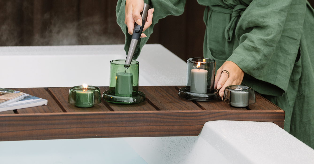 Person in green Citta robe lighting a candle on a wooden tray over an outdoor bathtub with three other candles and a magazine on the tray.