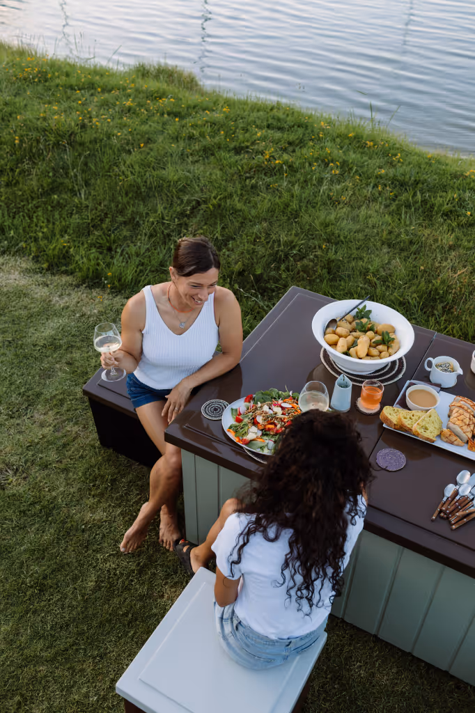Aerial view of two women sharing drinks beside a Star Baths outdoor bath with Eclipse Lid used as a dining table, Raglan New Zealand waterway