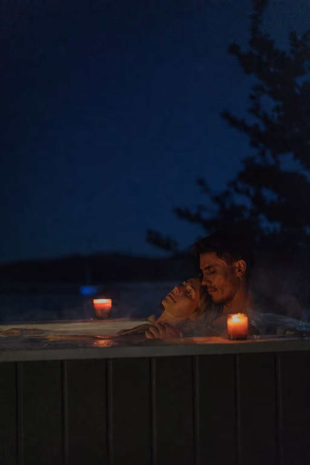 Couple embracing in a steaming Star Baths outdoor bath at night by candlelight with a dark blue sky and waterway silhouette