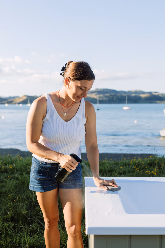 Woman cleaning a Star Baths outdoor bath edge with a spray bottle and sponge, Raglan harbour and sailboats in background