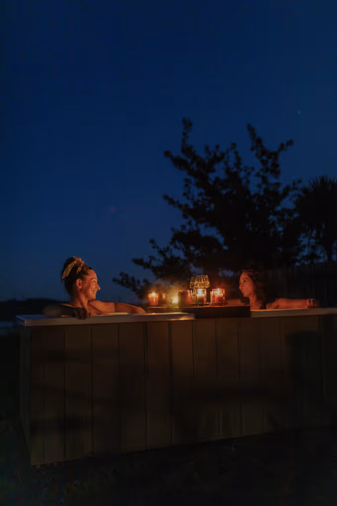 Two women laughing together in a Star Baths outdoor bath at night with candles and Retreat Tray, under a deep blue night sky