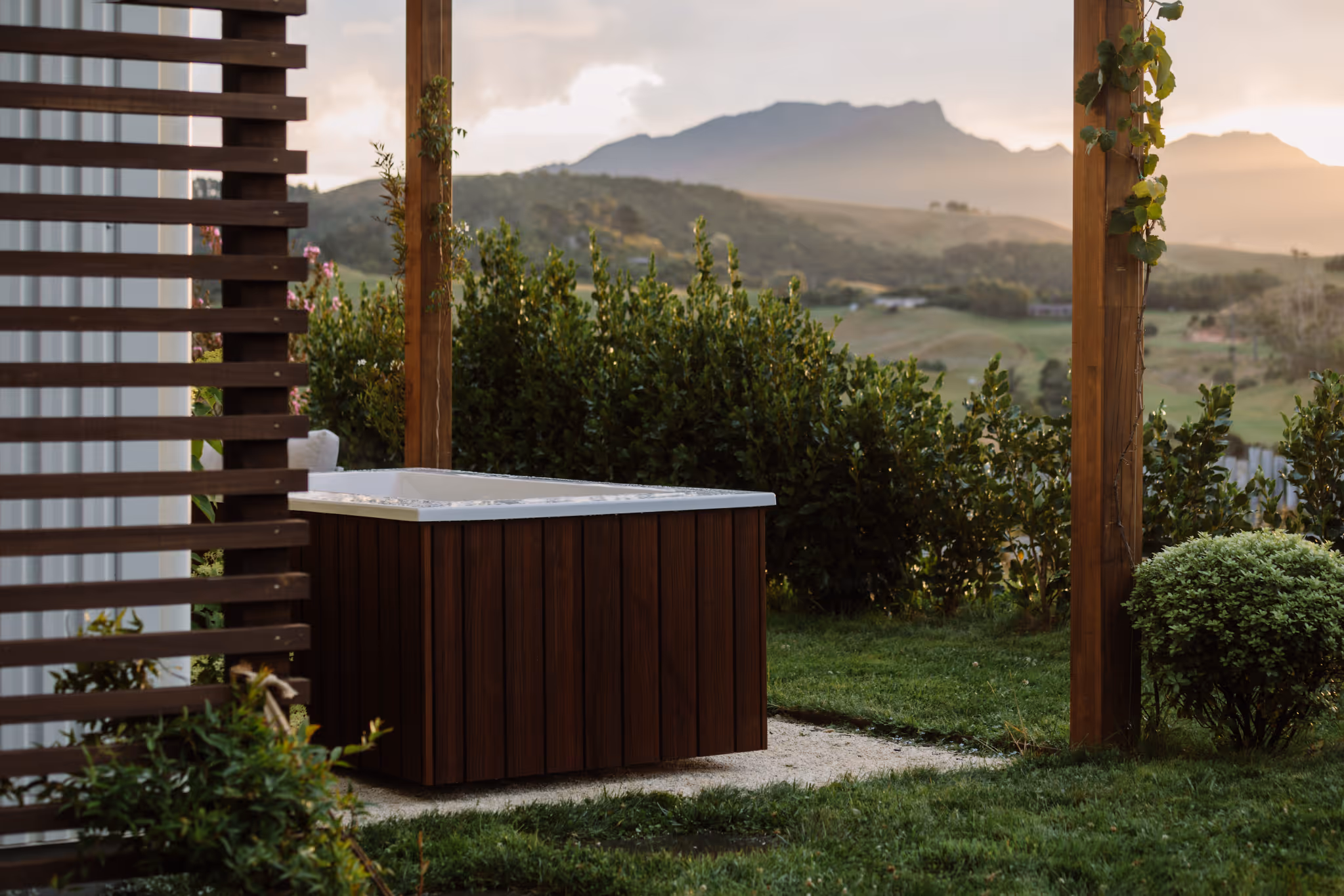Star Baths Signature Outdoor Bath with dark timber cladding set within a timber pergola garden, overlooking rolling green hills and mountains at dusk, New Zealand