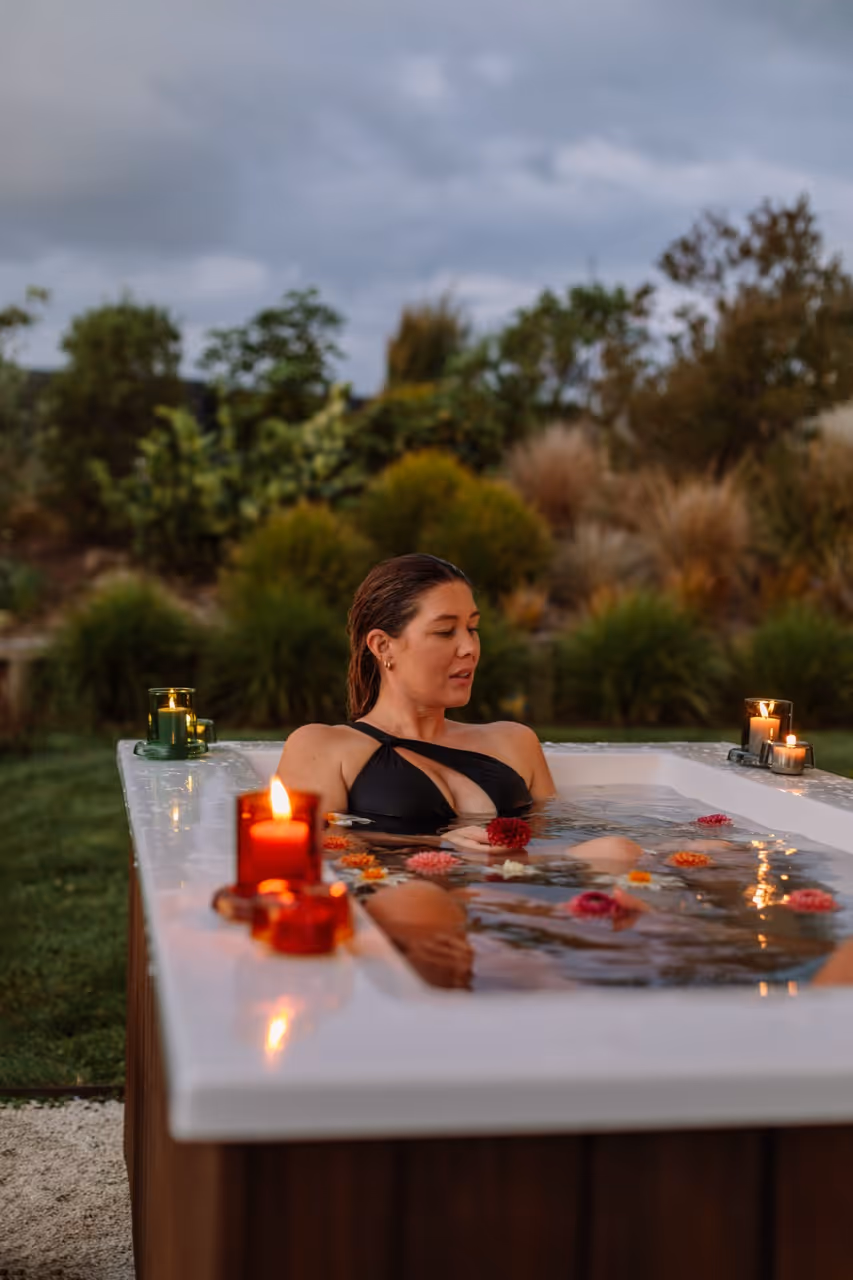 Woman relaxing alone in a Star Baths outdoor bath at dusk with floating flowers and candles on the wide edge, NZ garden backdrop