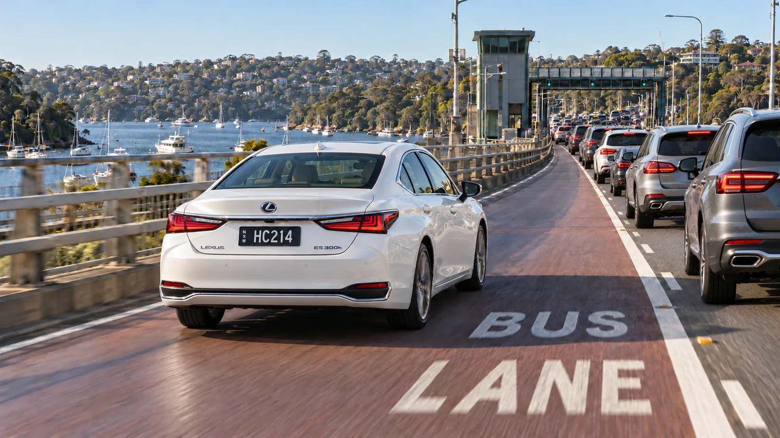 White Lexus ES 300h chauffeur car with NSW HC hire car plates driving in a red bus lane past queued traffic on a bridge over a Sydney harbour waterway