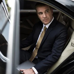 Businessman in a navy suit and gold tie seated in the leather rear seat of an executive car with a laptop, looking out of the open door