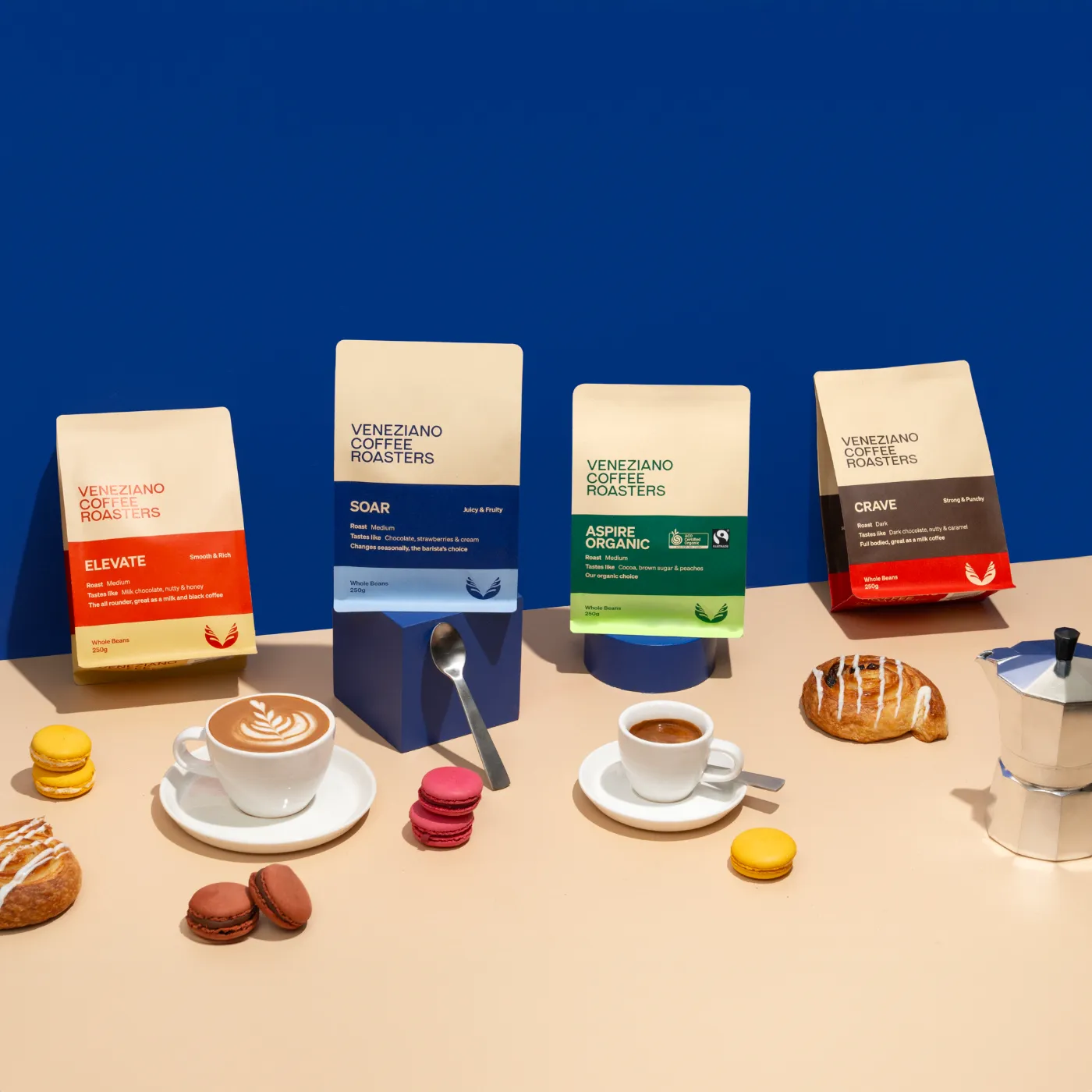 Stylised coffee product scene with Veneziano Coffee Roasters coffee bags, colourful macarons, pastries, and cups of coffee on a beige surface with a vibrant blue backdrop.