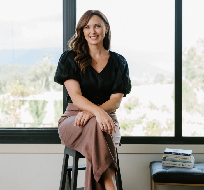 Lucy Quick, Founder of the Balanced Elite, sitting on a stool with a black note and beige pants.