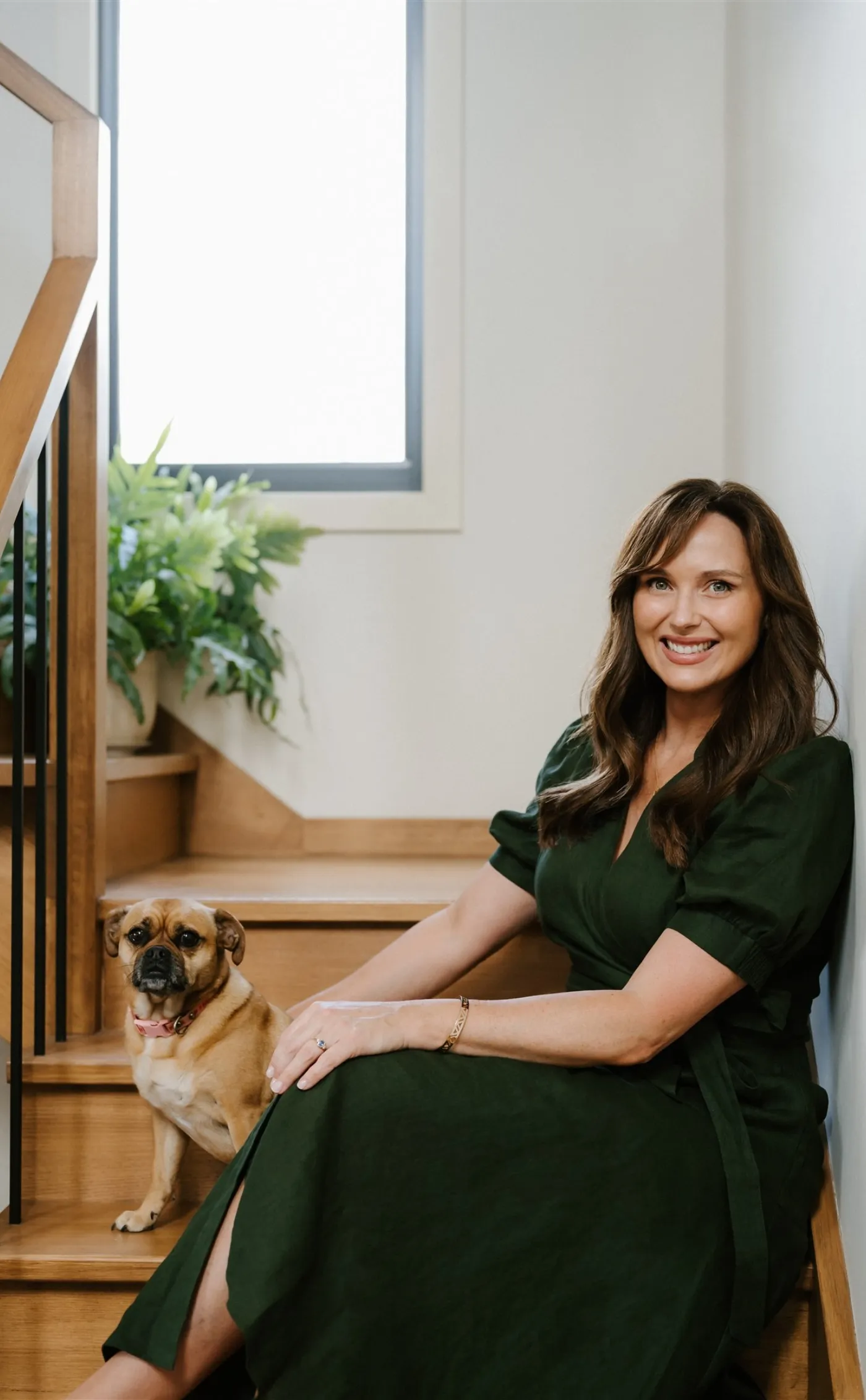 Lucy Quick, founder of The Balanced Elite, sitting on her stairs looking at the camera, in a deep green dress, with her dog at her side.