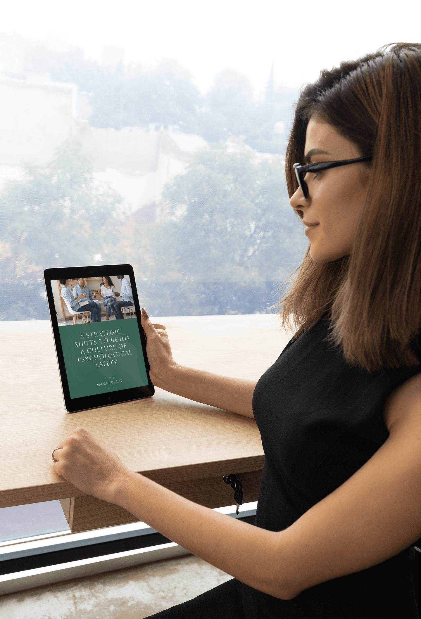A woman sitting at a cafe desk, holding her iPad with the eBook download of the '5 strategic shifts to build a culture of psychological safety'