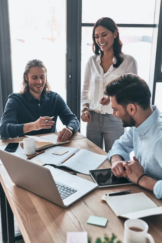 A woman standing over a desk with two men sitting at it, discussing happily about Imposter Syndrome and the tools that they're learning to combat it.