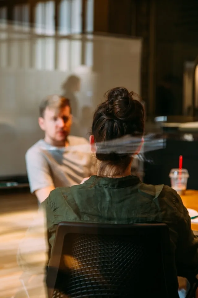 Two workplace colleagues facing each other behind a pane of glass in a boardroom, discussing work.
