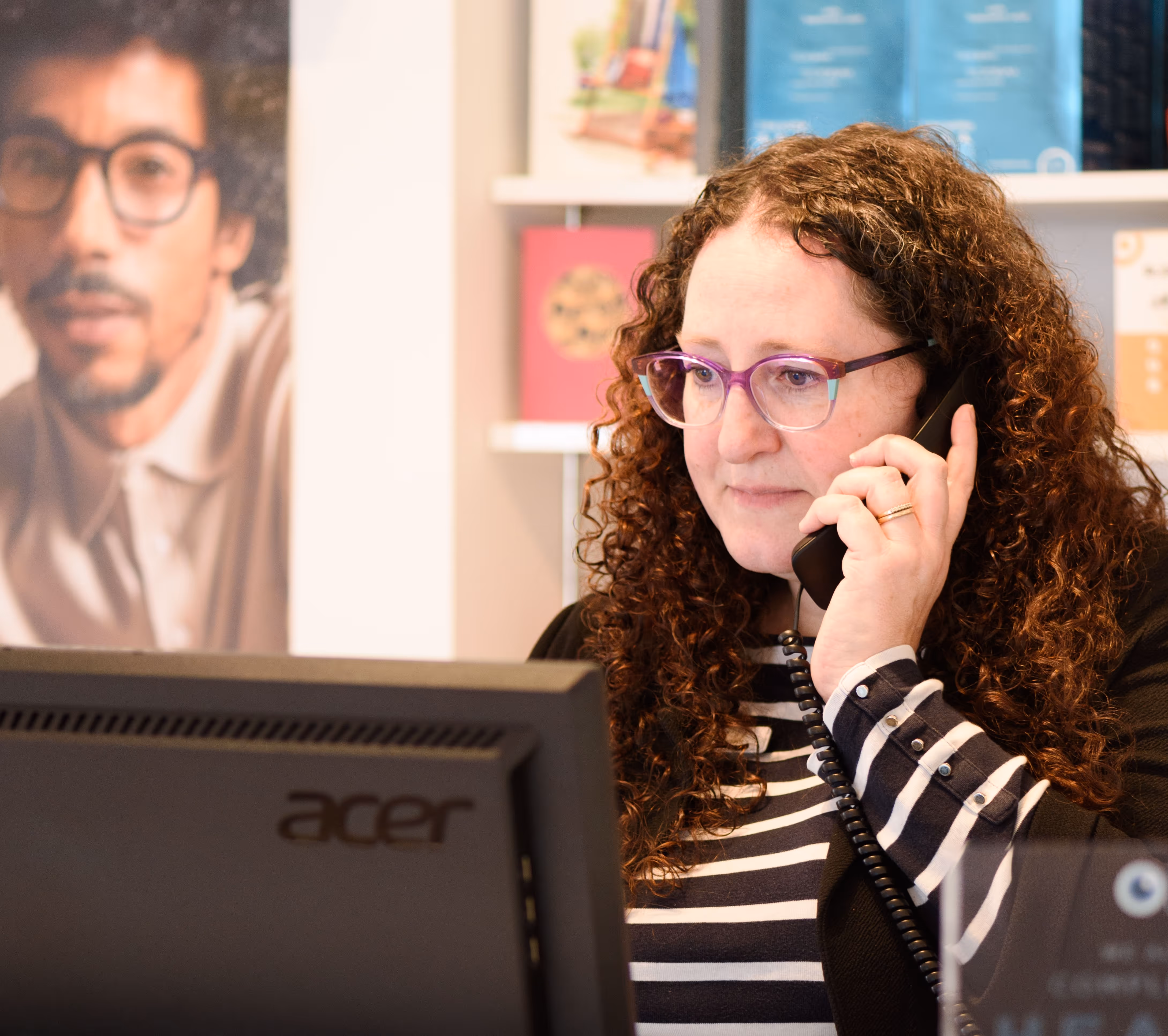 Woman with curly hair talking on phone near computer and poster