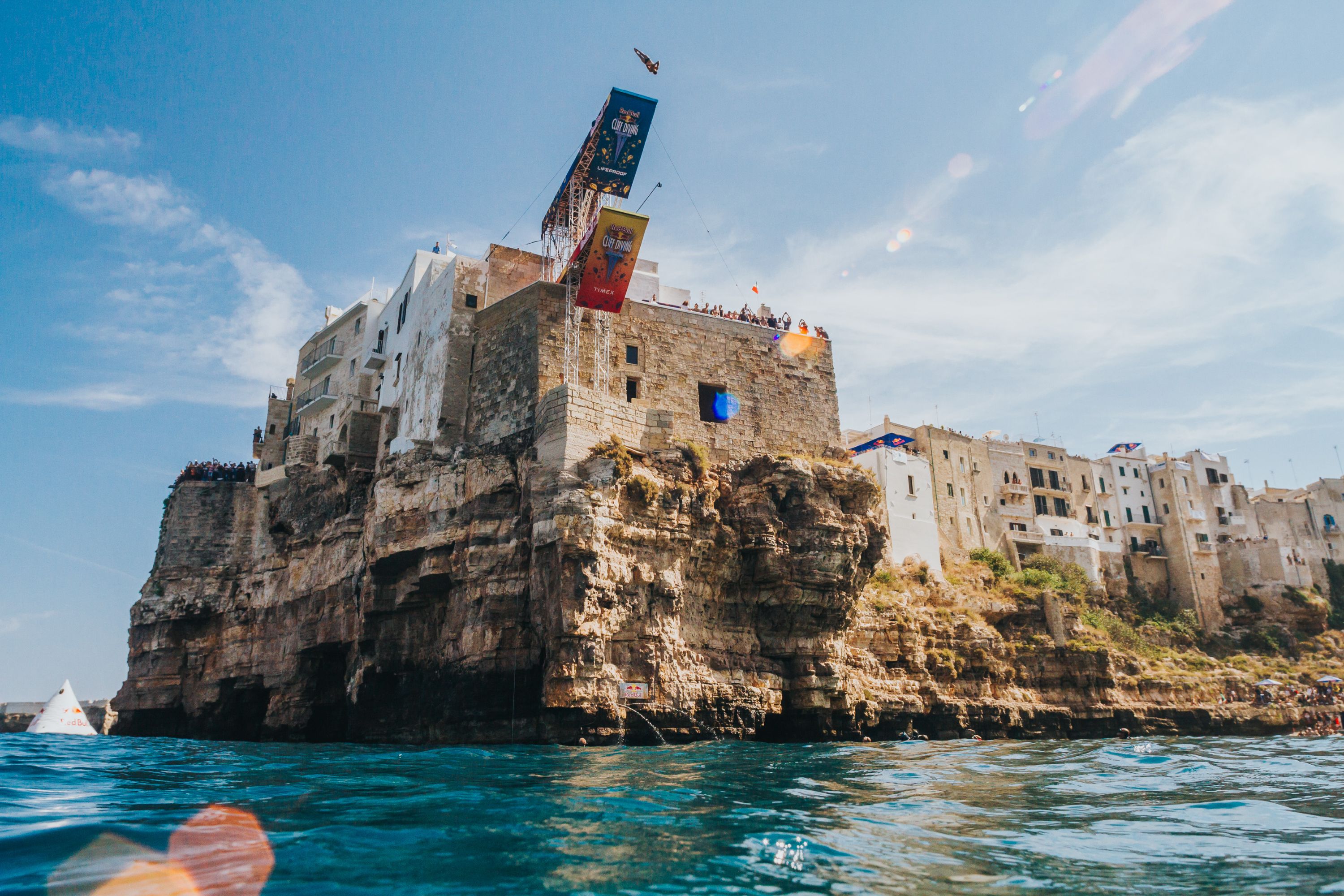A cliff, blue water and a person jumping to the water.