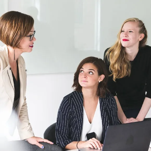 Three women engaged in a discussion around a laptop in a bright office setting.