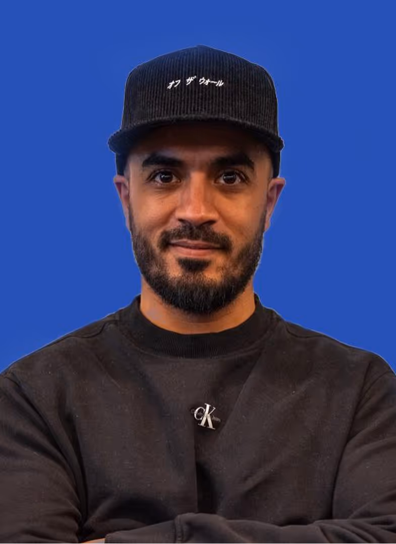 Homme portant une casquette noire avec des caractères japonais, un pull noir avec un logo CK, fond bleu uni.