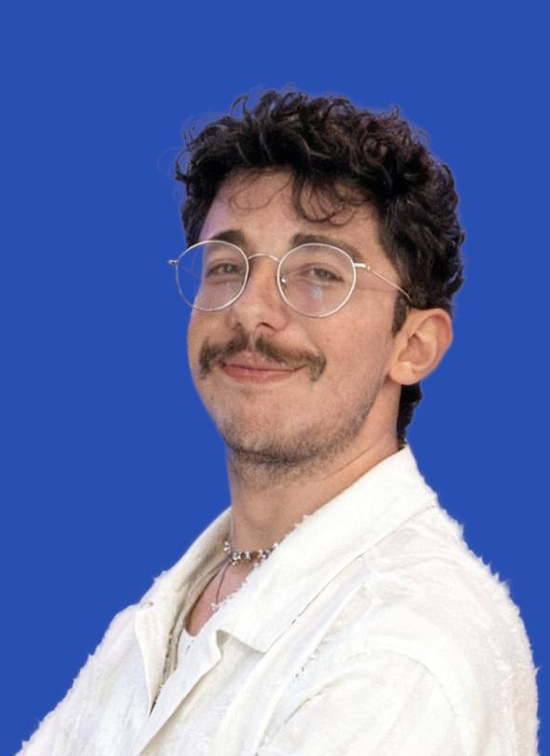 Jeune homme souriant avec lunettes rondes, moustache, cheveux bouclés et chemise blanche, fond bleu.