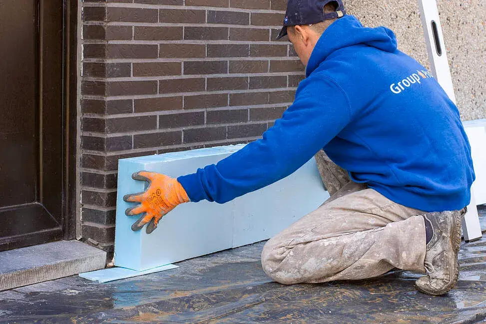 Bouwvakker in blauwe trui en werkbroek gebruikt een gele waterpas om blauwe XPS-isolatieplaten tegen een gevel recht te plaatsen, met handschoenen en werkschoenen.
