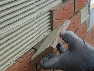 Close-up van een met handschoen gehulde hand die een steenstrip aanbrengt met lijm op een buitenmuur van baksteen.