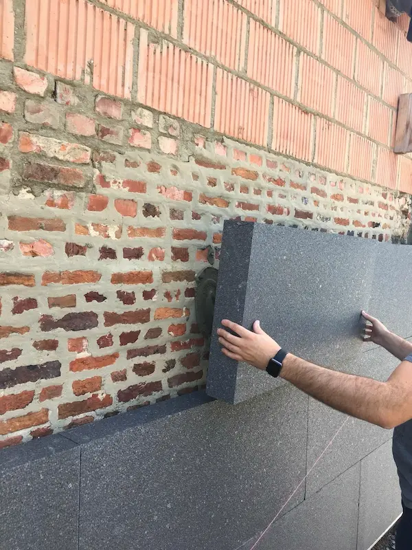 Man bevestigt donkergrijze isolatieplaat tegen buitenmuur van rode baksteen en snelbouwsteen, met roze aflijnkoord zichtbaar.