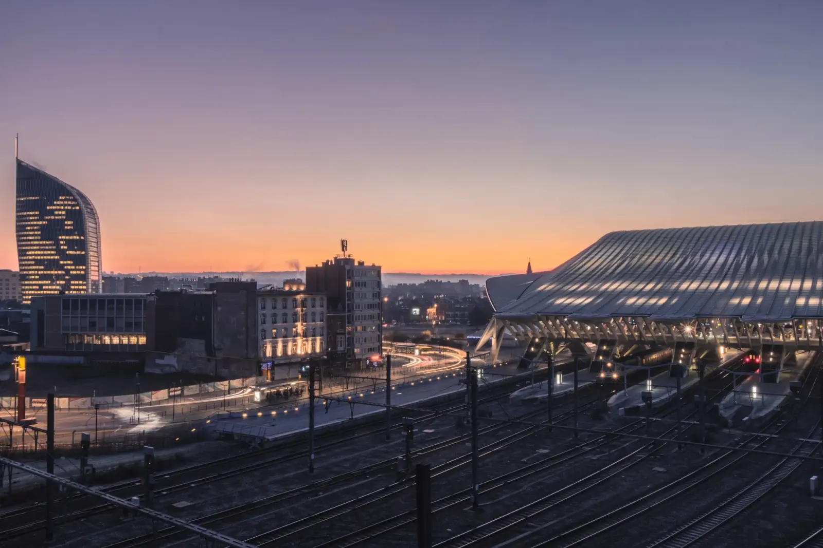 Avondzicht op Luik, België, met links de verlichte Tour Paradis wolkenkrabber en rechts het moderne station Liège-Guillemins, ontworpen door Santiago Calatrava, met treinsporen op de voorgrond en een oranje gekleurde zonsondergang.