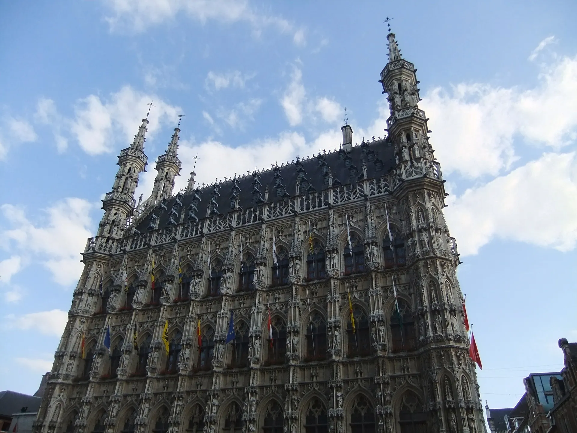 Het gotische stadhuis van Leuven, België, met rijk versierde gevel, talloze beelden en torentjes, gefotografeerd tegen een blauwe lucht met witte wolken.