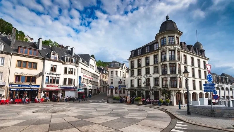 Gezellig stadsplein in de provincie Luxemburg, België, met cafés, terrasjes en het historische Hotel de la Poste onder een deels bewolkte hemel.
