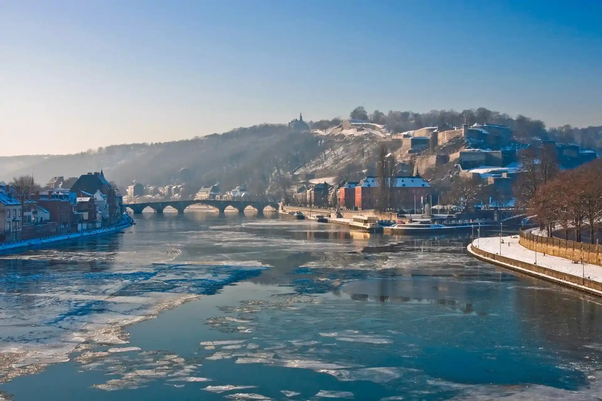 Winterzicht op de rivier de Samber in Namen, België, met drijvend ijs, kleurrijke gebouwen langs de oever, een stenen brug en de citadel van Namen op de heuvel.