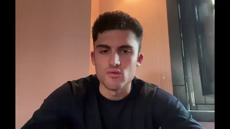 Young man with short curly hair wearing a black shirt sitting indoors near a window with blinds.