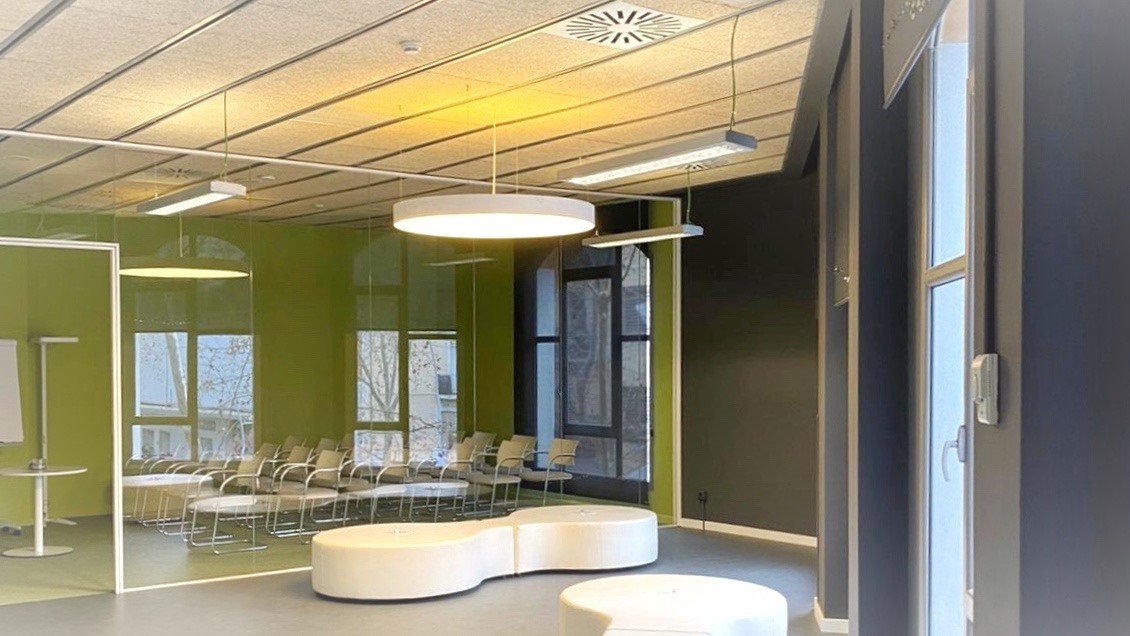 Sala riunioni moderna con sedie disposte in file dietro una parete di vetro, lampade sospese e pouf bianchi su pavimento grigio.