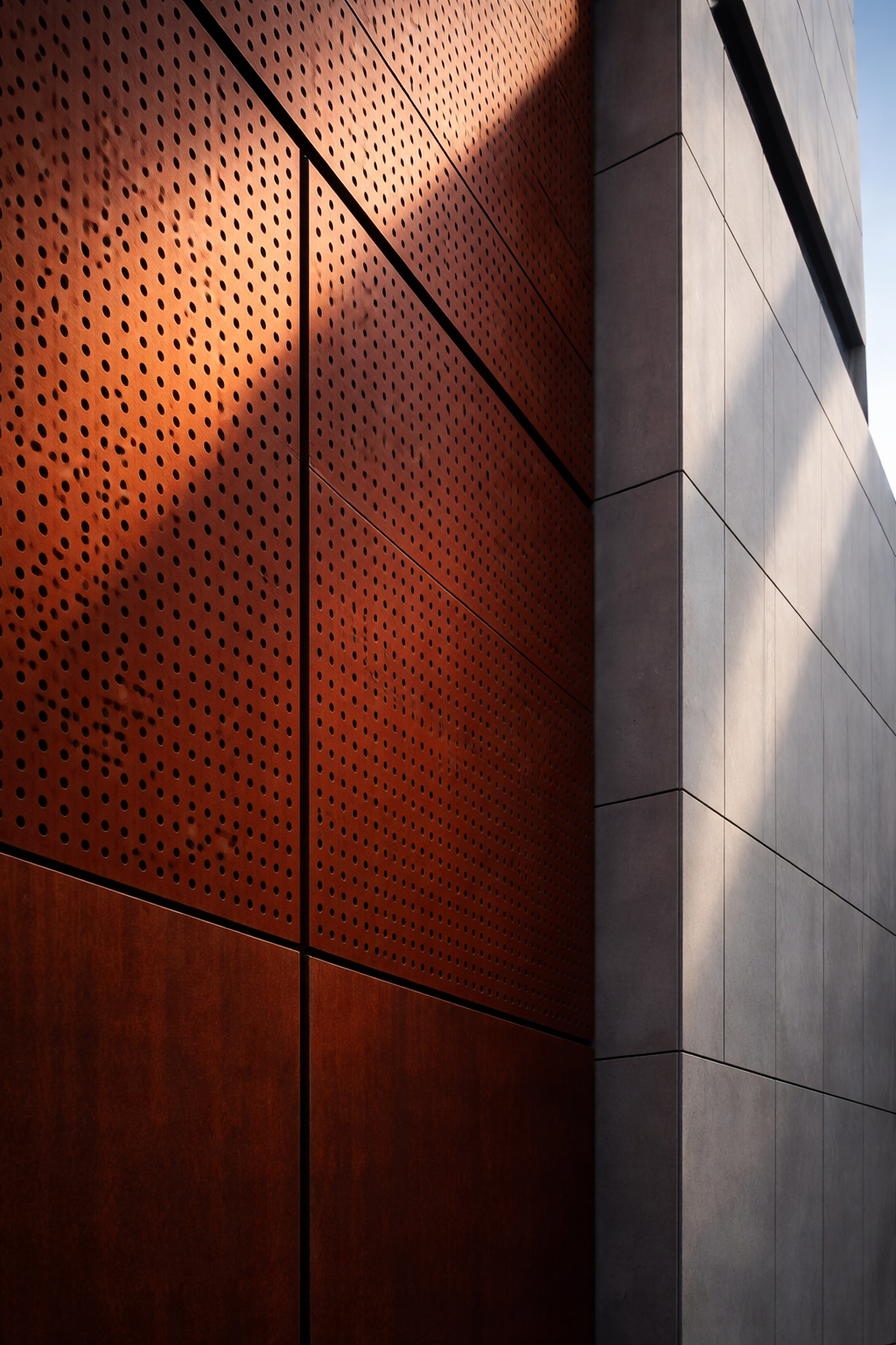 Particolare di una parete esterna con pannelli metallici ruggine forati e pannelli grigi lisci illuminati dal sole.